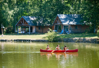 Kinder paddeln in einem Kanu auf einem See vor Holzhütten im Feriendorf Huttopia Lac de Rillé.