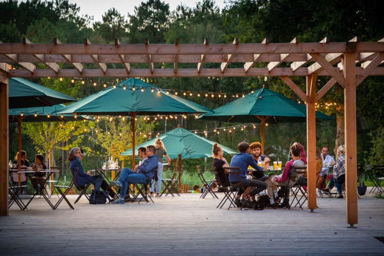 Outdoor dining area at Village Huttopia Lac de Rillé, guests seated under green umbrellas with string lights.
