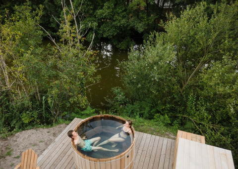Dos personas relajándose en una bañera al aire libre sobre una terraza de madera junto al lago en Huttopia Lac de Rillé.