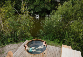 Twee personen genieten in een hottub op een houten terras vlak bij het water in Village Huttopia Lac de Rillé.
