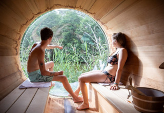 Dos personas en bañador se relajan en una sauna redonda con ventana grande y vistas a la naturaleza verde.
