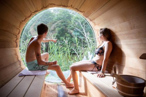 Dos personas en bañador se relajan en una sauna redonda con ventana grande y vistas a la naturaleza verde.