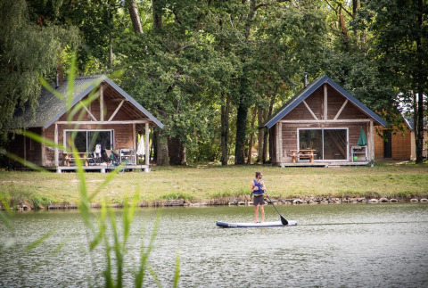Persona practicando paddle surf en un lago frente a dos cabañas de madera rodeadas de bosque en Francia.