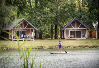 En person står på paddleboard på en sø foran to hyggelige træhytter omgivet af grøn skov i Frankrig.