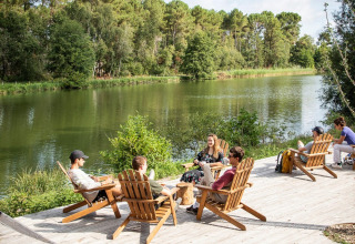 Mensen zitten ontspannen in houten zetels aan een meer in Village Huttopia Lac de Rillé, omgeven door bos.