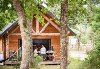 Familie entspannt vor einer Holzhütte im Grünen im Village Huttopia Lac de Rillé, Centre-Val de Loire.