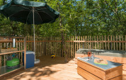 Outdoor deck at a teepee tent with grill, umbrella, wooden fence, seating, and lush forest scenery.