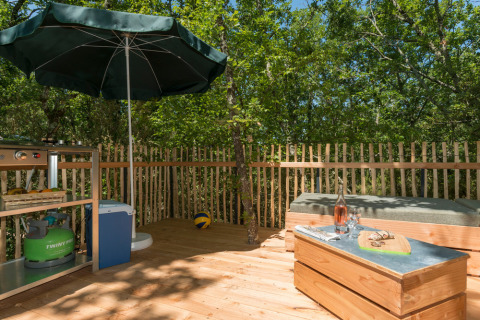 Buitenterras aan een tipi-tent met barbecue, parasol, houten omheining, zitbank en groene omgeving.