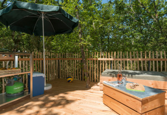 Terrasse extérieure à une tente tipi avec barbecue, parasol, banc, table et environnement boisé.