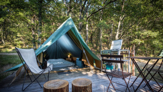 Teepee-telt i skoven med træstole, klapstole og campingkogeplade på en træterrasse.
