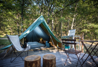 Teepee tent in a forest with wooden stools, camping chairs, and a small kitchen on a wooden deck.