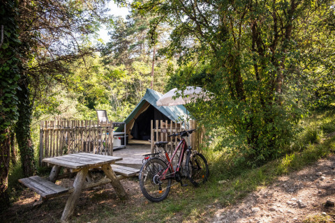 Emplacement de camping avec tente tipi, vélo, table en bois et arbres verts en pleine nature.