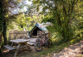 Área de camping con tienda tipi, bicicleta y mesa de madera, rodeada de árboles y naturaleza verde.