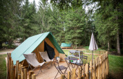 Una tienda tipi sobre una terraza de madera en un bosque, con sillas, mesa, parrilla y cerca rústica.