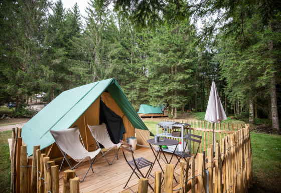 Een tipi-tent op een houten platform in het bos met stoelen, tafel, barbecue en een omheind tuintje.