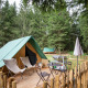 En tipi-telt på en træterrasse omgivet af skov, med stole, bord og grill i et hyggeligt naturområde.