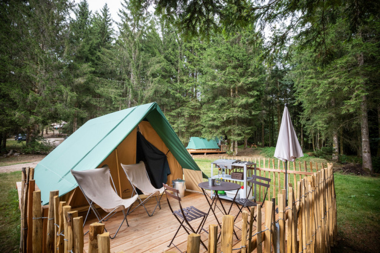 A teepee tent on a wooden deck in the forest, featuring chairs, table, grill, and a rustic fenced area.