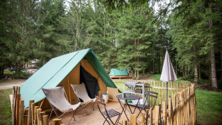 Una tienda tipi sobre una terraza de madera en un bosque, con sillas, mesa, parrilla y cerca rústica.