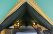 Interior de una tienda tipi con cama, mesitas, lámparas y bolsas, mostrando un ambiente acogedor.