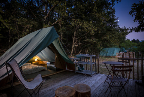 Foto nocturna de un campamento con tienda tipi, asientos exteriores y luz encendida en el bosque.