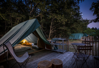 Evening photo of a teepee tent campsite with outdoor seating and lamp glowing inside in the forest.