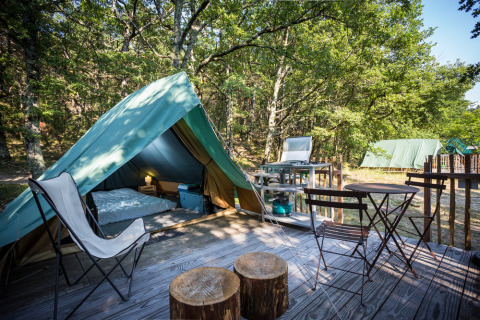 Tienda tipi sobre plataforma de madera con sillas, mesa y cocina de camping rodeada de árboles.