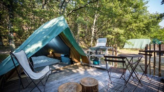 Tienda tipi sobre plataforma de madera con sillas, mesa y cocina de camping rodeada de árboles.