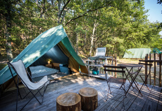 Tienda tipi sobre plataforma de madera con sillas, mesa y cocina de camping rodeada de árboles.