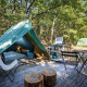 Tienda tipi sobre plataforma de madera con sillas, mesa y cocina de camping rodeada de árboles.