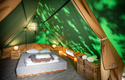 Vista interior de una tienda tipi con cama doble, mesitas de noche, lámparas y cesta con toallas.