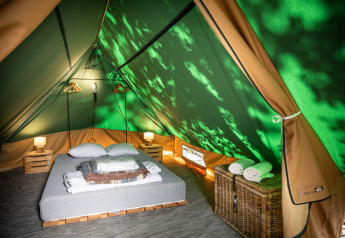 Vista interior de una tienda tipi con cama doble, mesitas de noche, lámparas y cesta con toallas.