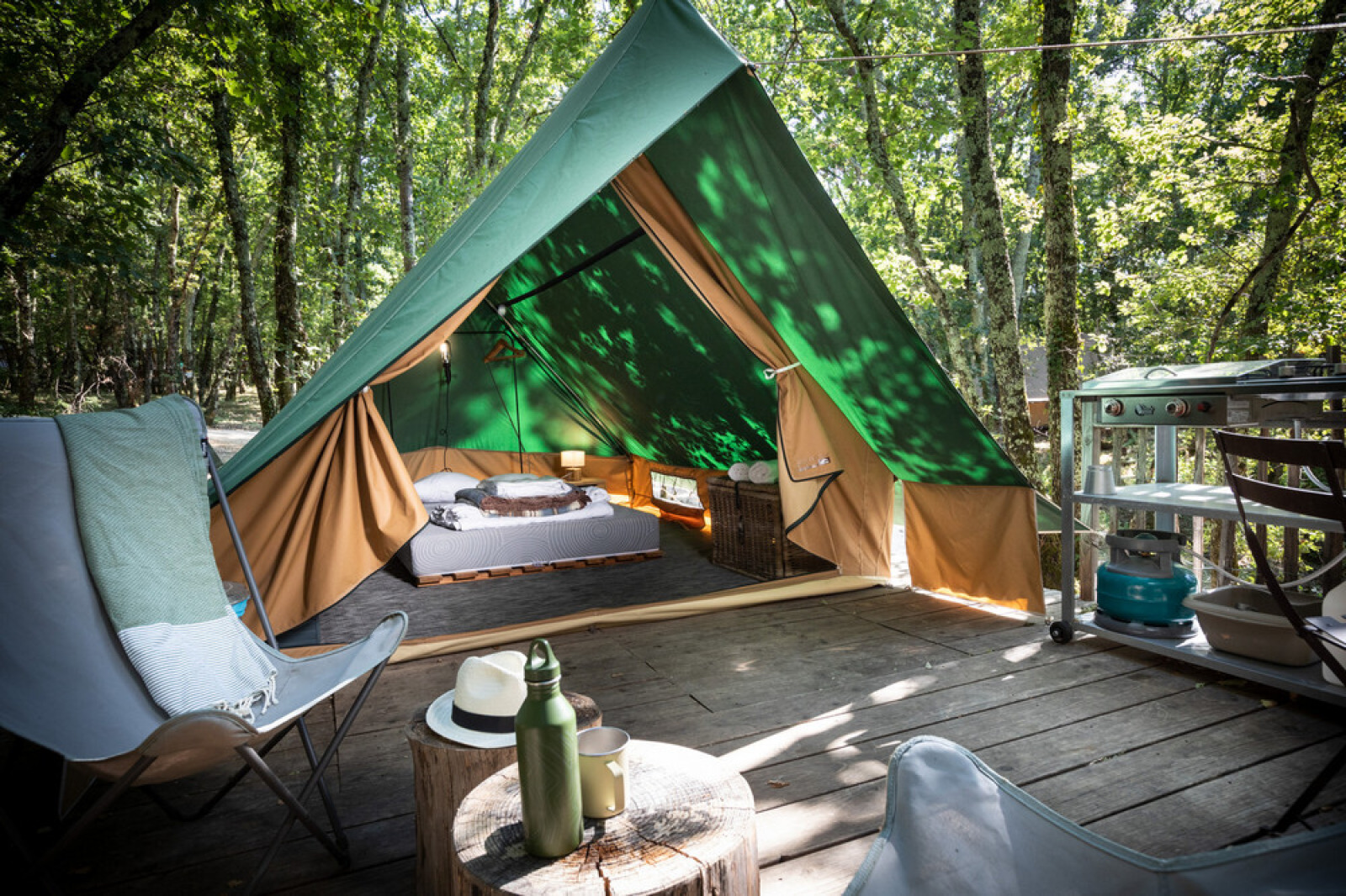 Tipitent op een houten terras in het bos, met een gezellig bed, stoelen en een kleine buitenkeuken.
