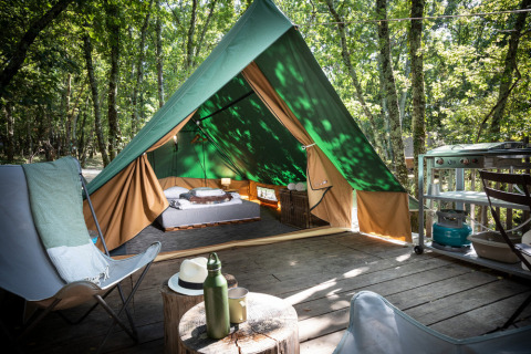 Una tienda tipi sobre una tarima de madera en el bosque, equipada con cama, sillas y pequeña cocina exterior.