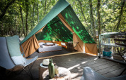 Une tente tipi sur une terrasse en bois dans la forêt, aménagée avec un lit, des chaises et une cuisine extérieure.