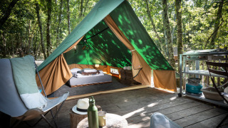 Una tienda tipi sobre una tarima de madera en el bosque, equipada con cama, sillas y pequeña cocina exterior.