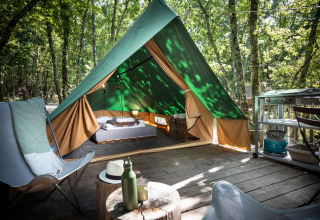 Una tienda tipi sobre una tarima de madera en el bosque, equipada con cama, sillas y pequeña cocina exterior.
