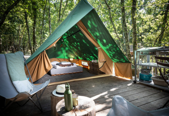 Una tienda tipi sobre una tarima de madera en el bosque, equipada con cama, sillas y pequeña cocina exterior.