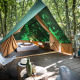 En teepee-telt på en træterrasse i en skov, hyggeligt indrettet med seng, stole og et lille udendørskøkken.