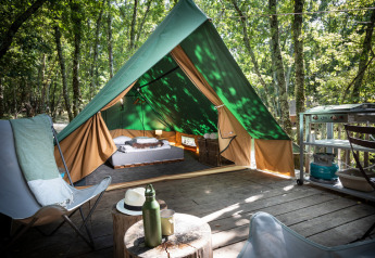 Una tienda tipi sobre una tarima de madera en el bosque, equipada con cama, sillas y pequeña cocina exterior.