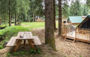 Billede af et tipitelt på en træterrasse i en skov med bænk og bæk, ideelt til glamping.