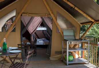 Tente safari avec terrasse, table extérieure, barbecue et vue sur la forêt. Hébergement glamping moderne.