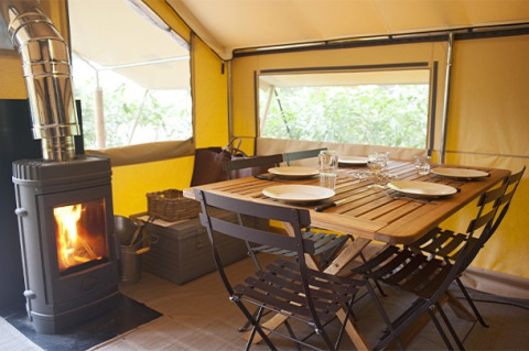Interior de una tienda safari con mesa de madera puesta para cuatro y estufa de leña encendida junto a la mesa.
