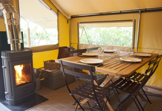 Interior de una tienda safari con mesa de madera puesta para cuatro y estufa de leña encendida junto a la mesa.