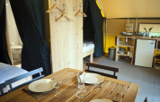 Interior view of a safari tent featuring a wooden dining table, kitchen area, beds, and a bathroom.