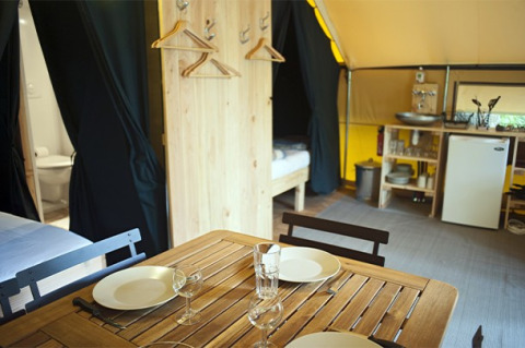 Vista interna di una tenda safari con tavolo da pranzo, angolo cucina, letti e bagno visibile.