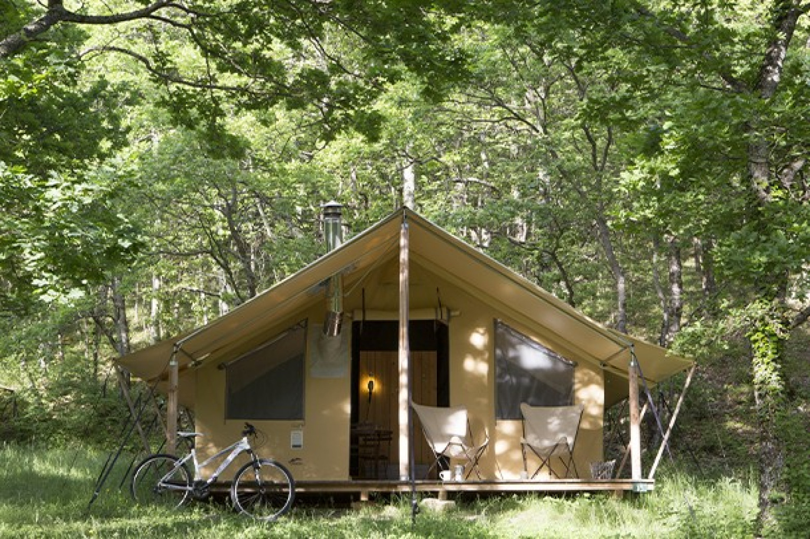 Safaritent in het bos met twee stoelen en een fiets ervoor, omringd door groene bomen en zonlicht.