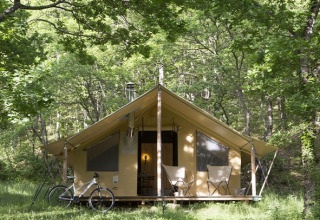 Tienda safari en el bosque con dos sillas y una bicicleta fuera, rodeada de árboles verdes y naturaleza.