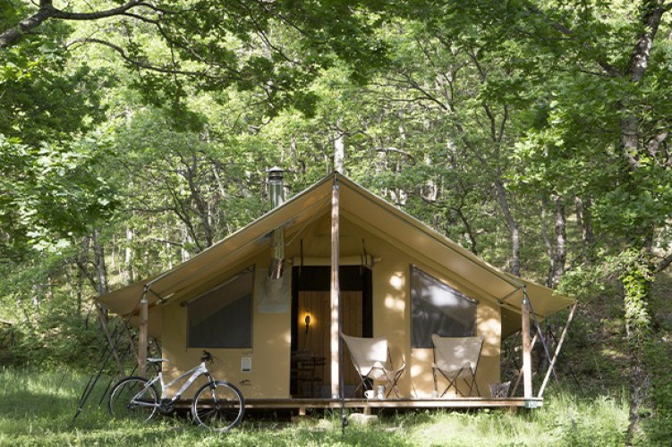 Tienda safari en el bosque con dos sillas y una bicicleta fuera, rodeada de árboles verdes y naturaleza.