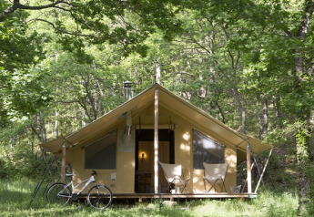 Safaritent in het bos met twee stoelen en een fiets ervoor, omringd door groene bomen en zonlicht.
