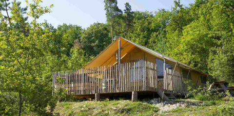 Safari tent with wooden deck and fence, nestled among green trees in a peaceful, forested landscape.